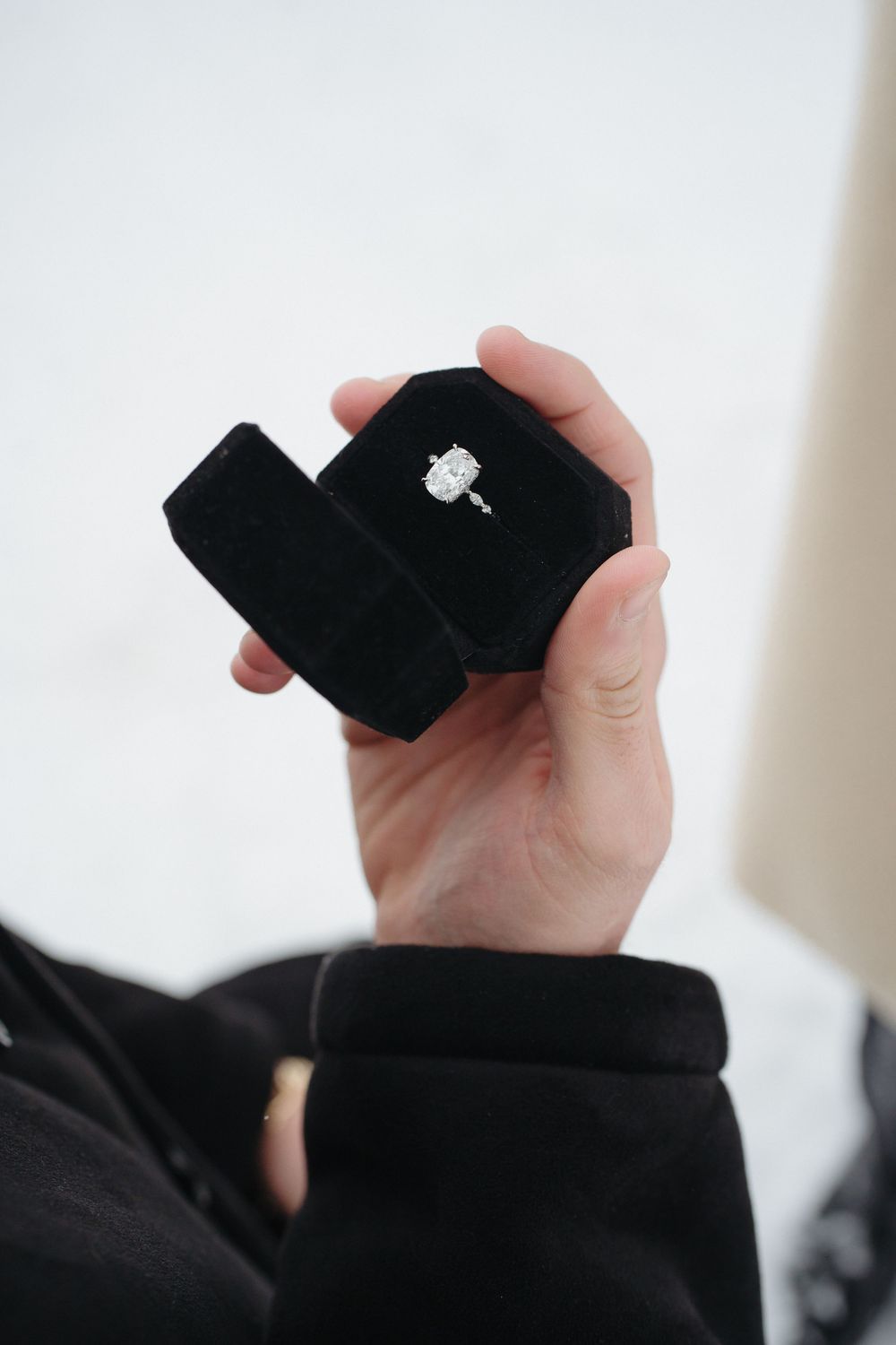 A black ring box being held open to display a sparkling diamond engagement ring.