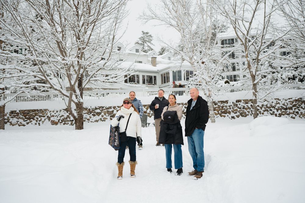Three people stand together in a snowy winter landscape with a white house and snow-covered trees in the background.