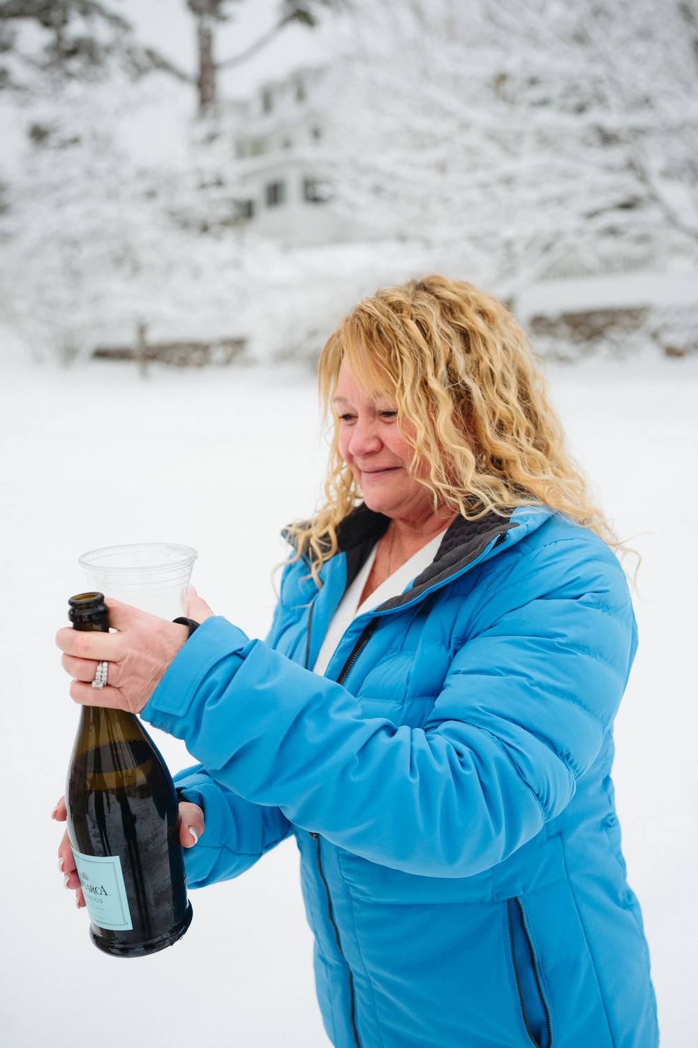 Person in bright blue winter coat opening a champagne bottle in snowy outdoor setting.