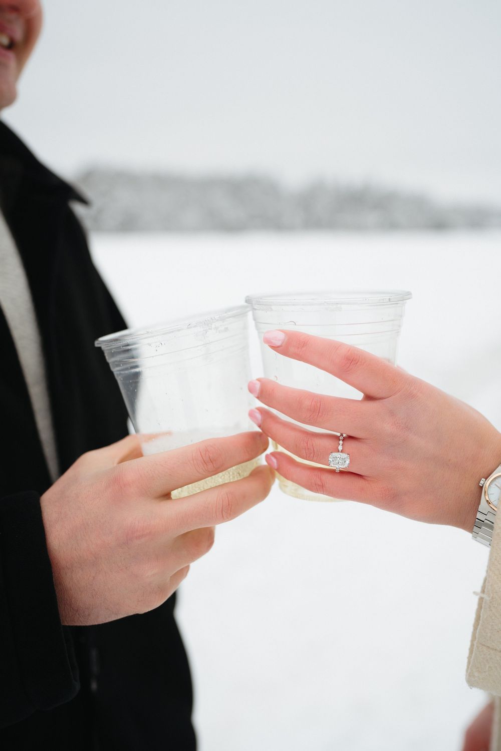 Two hands hold clear plastic cups in a winter setting with snow in the background.