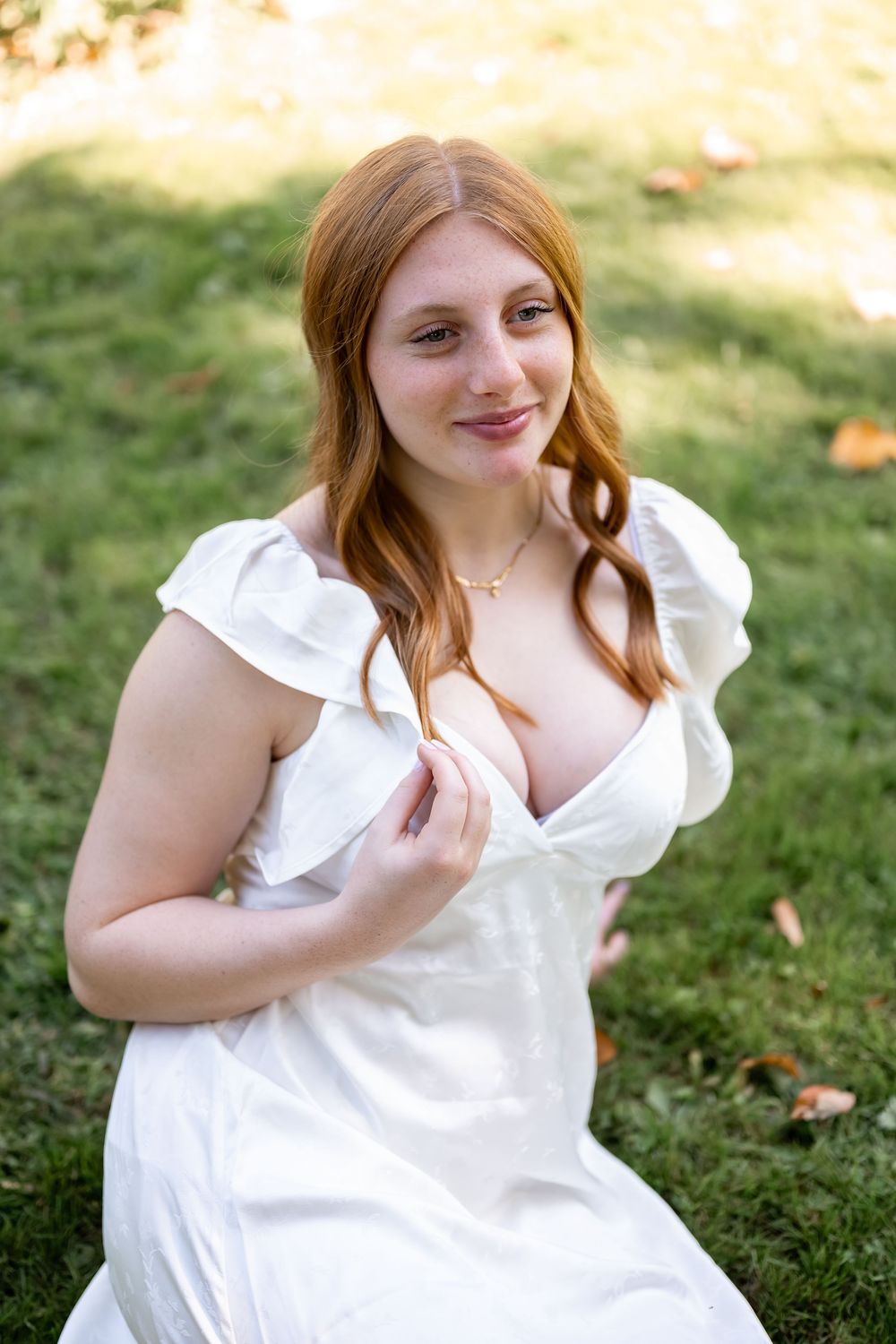 A person in a white ruffled dress sits on grass in a sunny outdoor setting with autumn leaves scattered around.