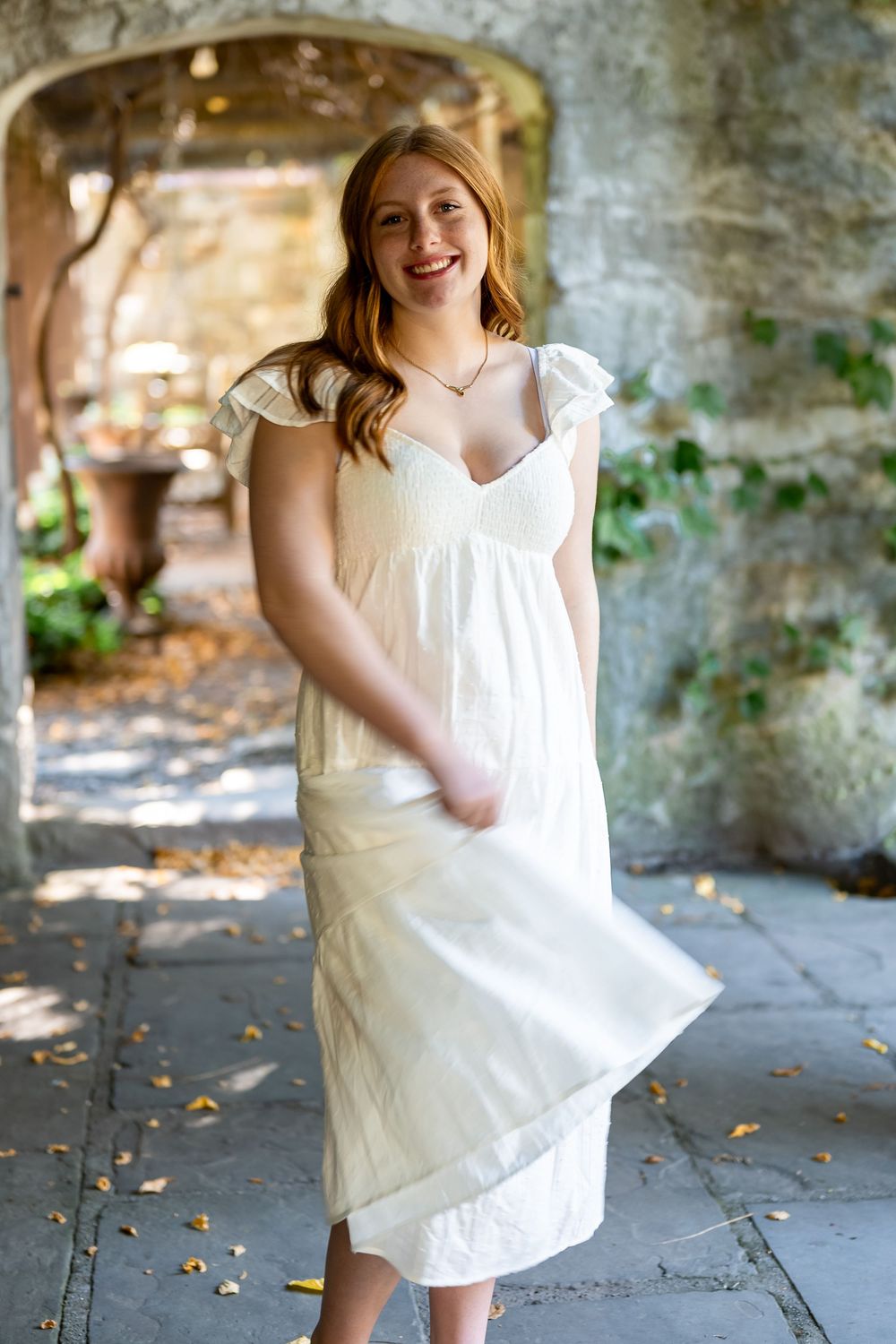 A person in a flowing white summer dress twirls and dances joyfully in a stone archway on a sunny day.