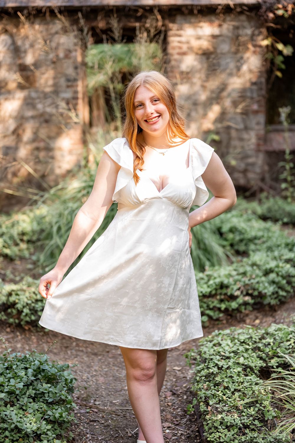 A white summer dress twirls in a garden setting with stone walls and green foliage in the background.