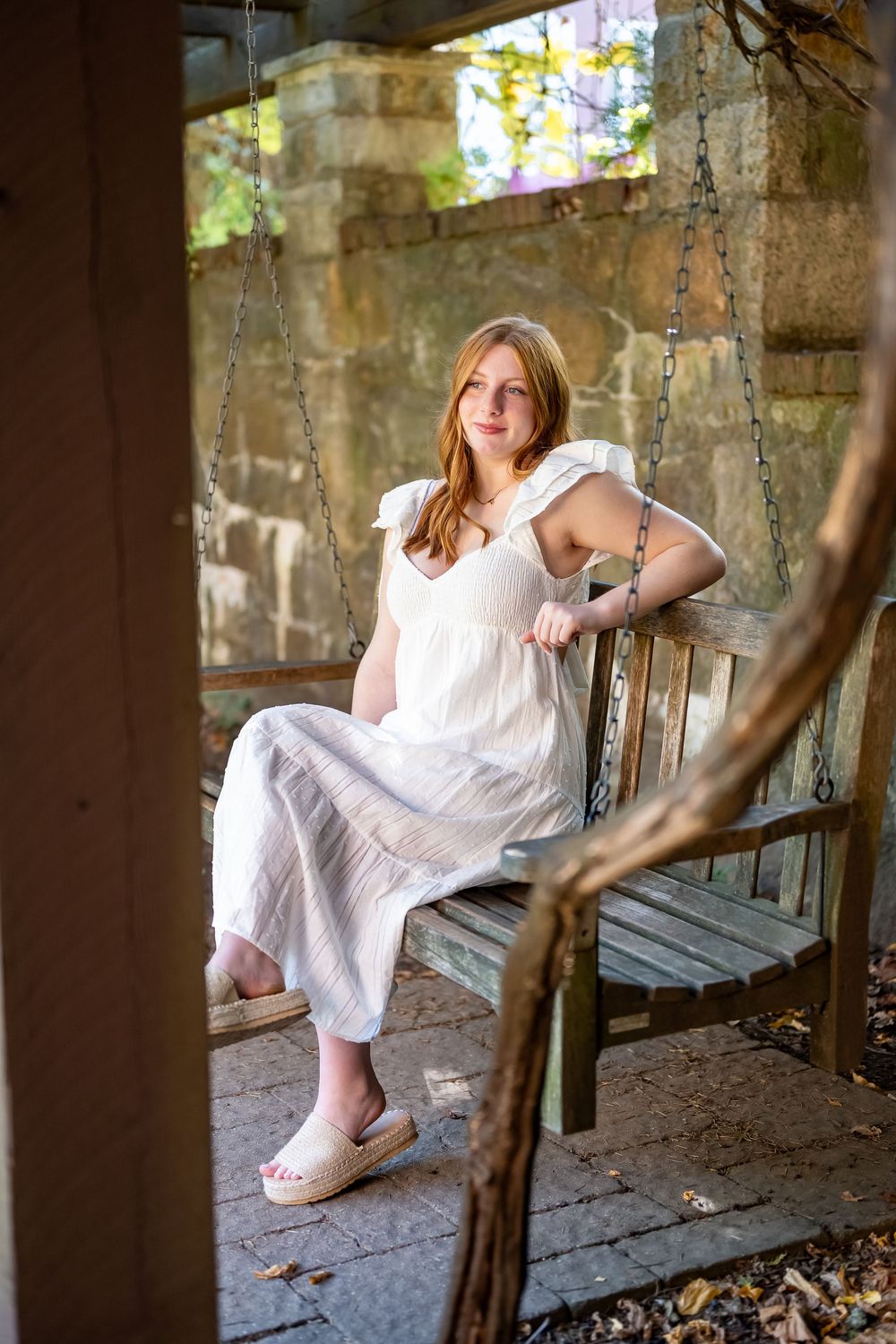A person in a white dress sits on a rustic wooden bench in dappled sunlight surrounded by stone walls.