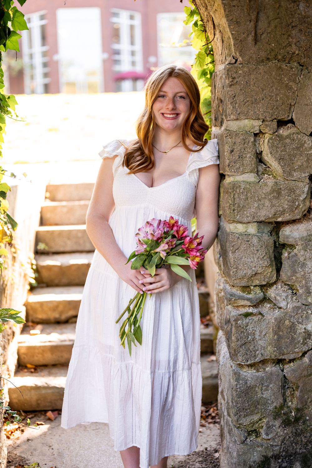 A person in a white dress holds a colorful bouquet while standing near stone stairs in an outdoor setting.