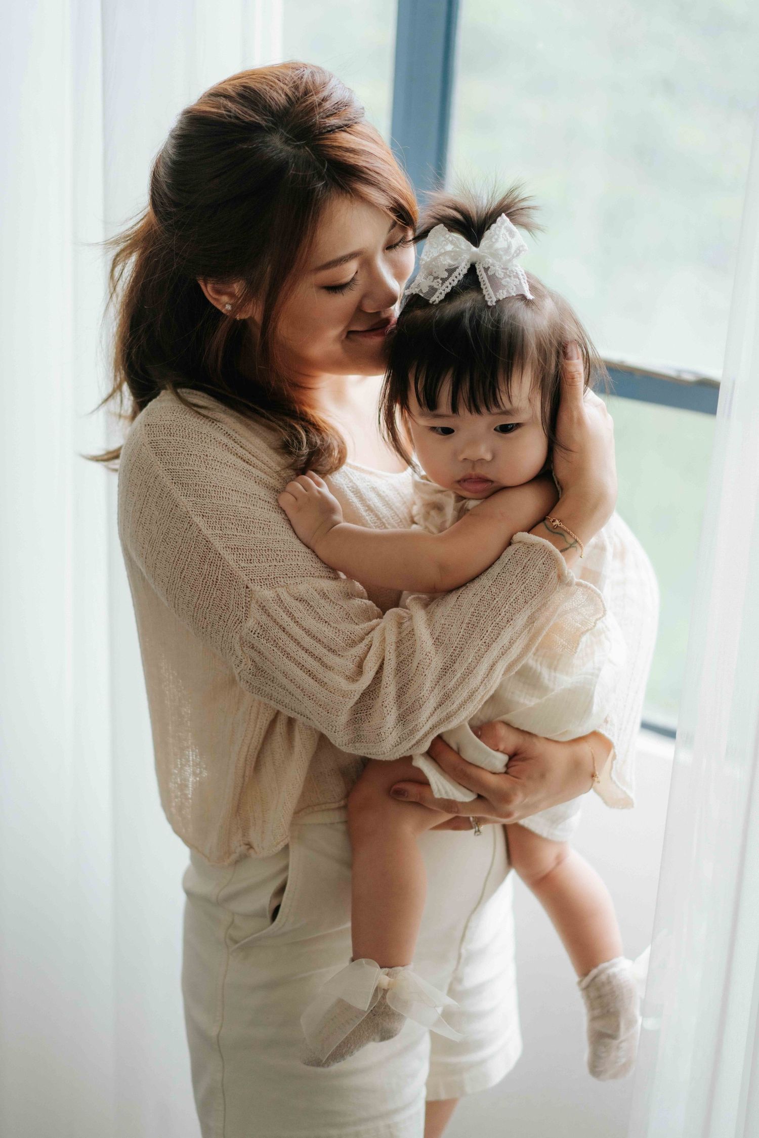 A parent in white clothing holds and embraces a baby by a bright window in a tender moment一位身穿白色衣服的家長在明亮的窗邊抱著嬰兒，畫面溫馨感人。