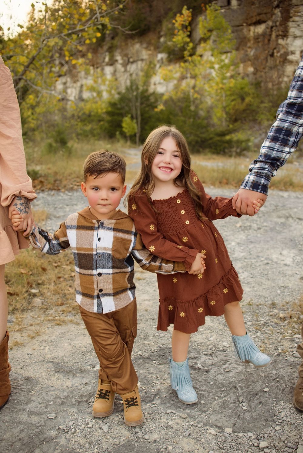 Family members in coordinated brown and plaid outfits hold hands while walking on a gravel path in autumn.