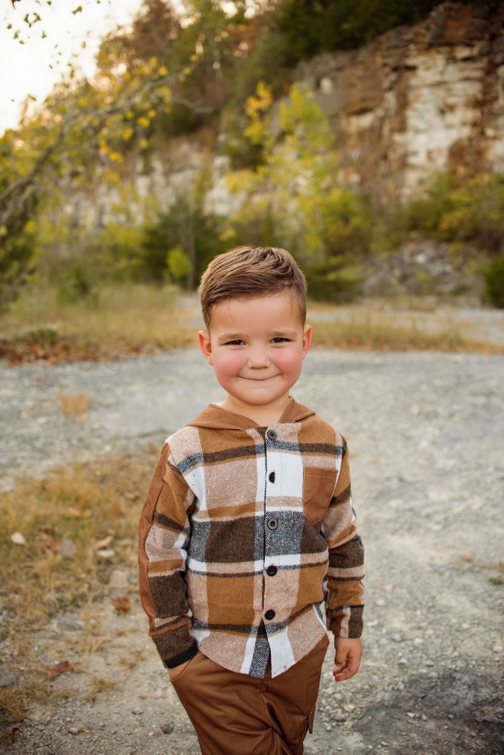 A young child in a brown plaid sweater poses outdoors against a rocky natural backdrop on an autumn day.