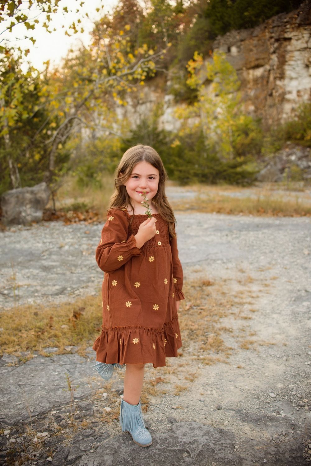 Child wearing brown fringe suede jacket and jeans poses outdoors against rocky backdrop in autumn setting.