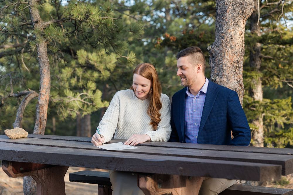 A couple sits together at a rustic wooden picnic table in a sunny outdoor forest setting.