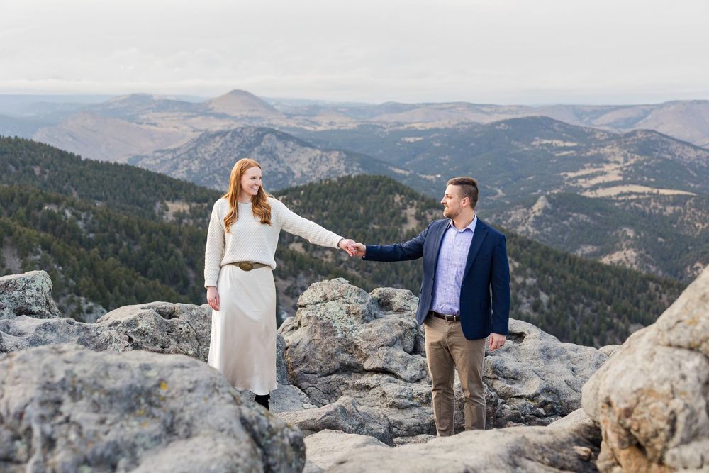 Two people sit together on a scenic mountain cliff edge overlooking a vast mountain landscape.