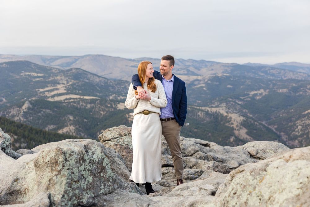A couple shares a romantic moment on a mountain peak overlooking a scenic valley landscape.