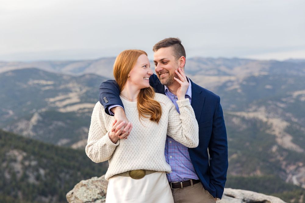 A couple embraces while standing on rocky mountain terrain with sweeping mountain views in the background.