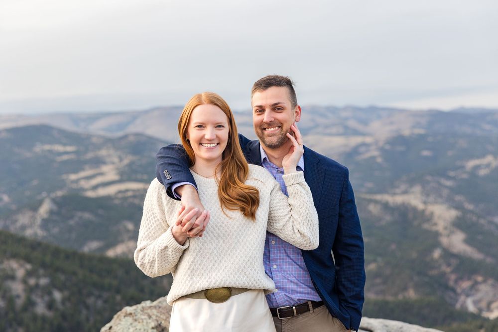 A couple smiles together on a mountain summit with panoramic views of rolling hills behind them.