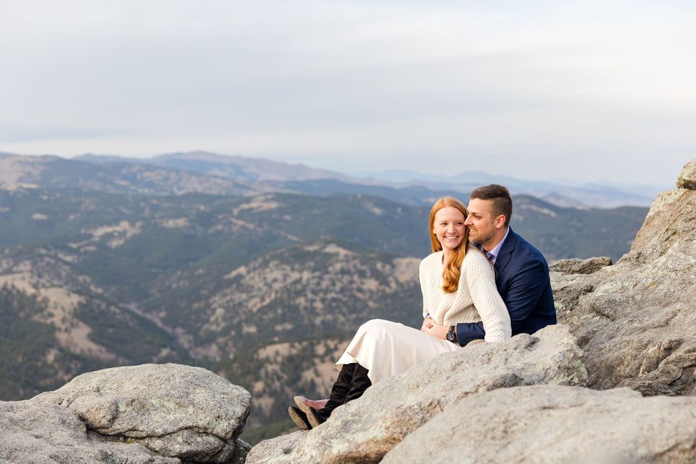 Couple sits on rocky mountain ledge overlooking vast mountain landscape on sunny day.
