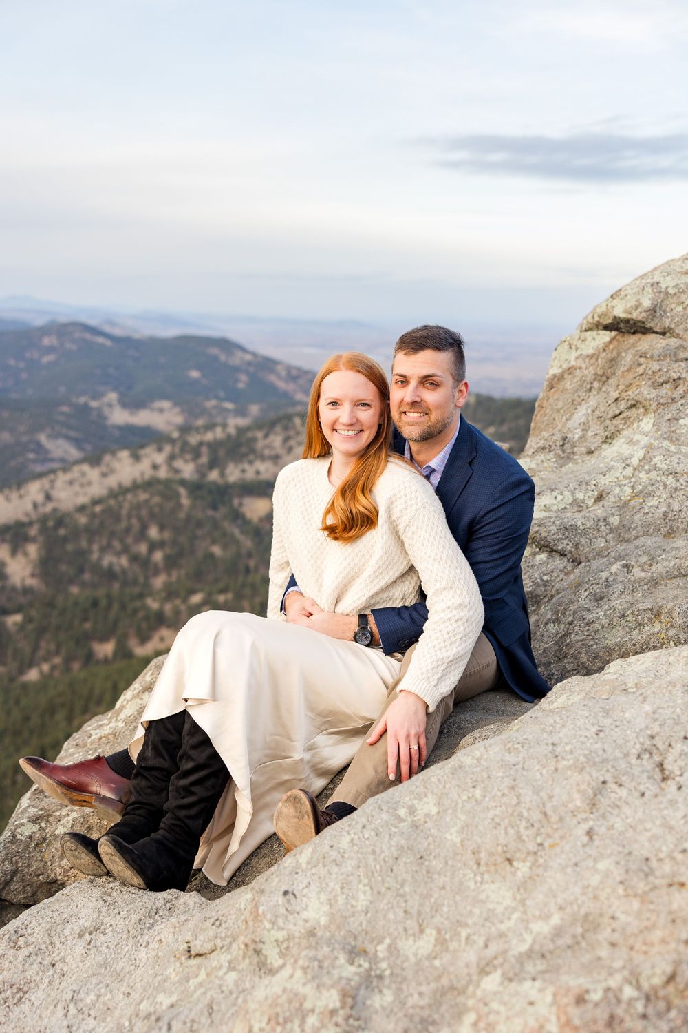 A couple sits together on rocky mountain ledge overlooking scenic mountain vista during engagement photo session.