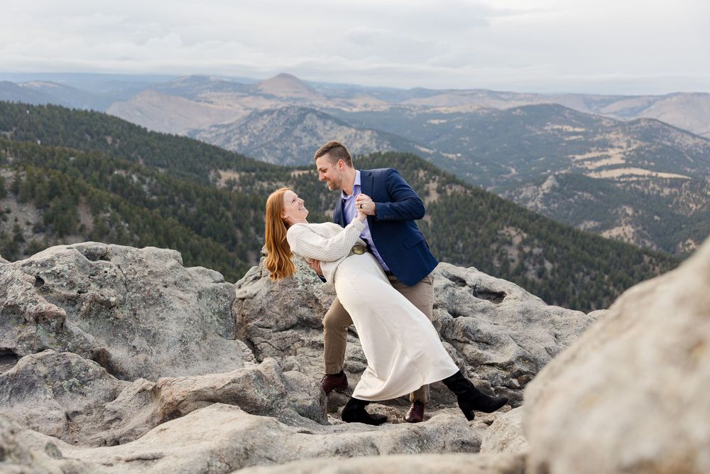 A couple shares an intimate moment on a scenic mountain overlook with dramatic rocky terrain and distant peaks.