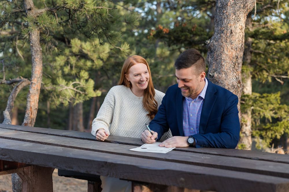 A couple sits together on a rustic wooden bench in a sunlit forest setting during a romantic photoshoot.