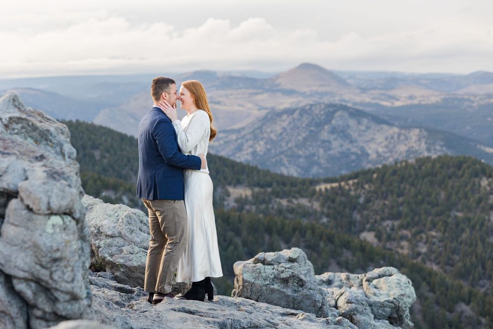 A couple shares a romantic moment on a mountain peak overlooking a scenic valley landscape at sunset.