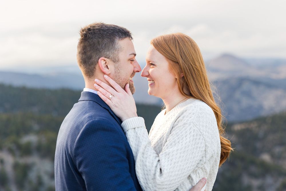 A romantic couple shares an intimate moment while standing on a mountain overlook with scenic views in the background.