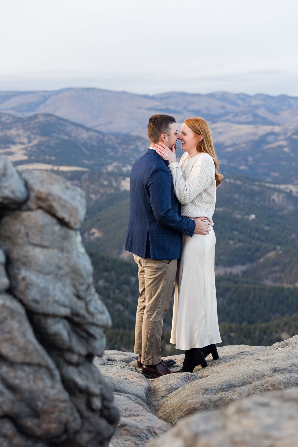 A couple shares a romantic moment atop a scenic mountain overlook with sweeping valley views in the background.