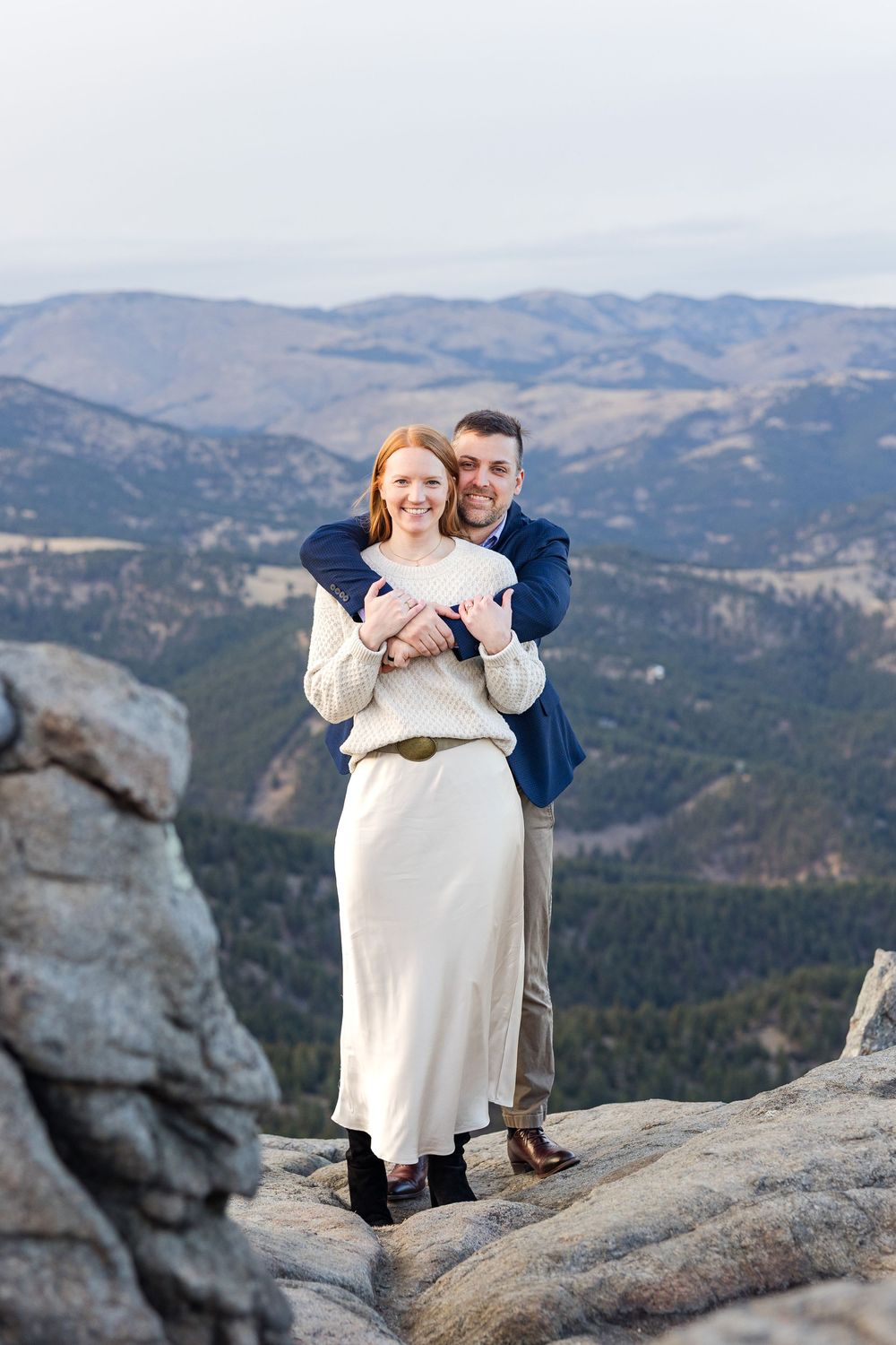 A couple shares a romantic moment on a rocky mountain overlook with sweeping mountain vistas in the background.