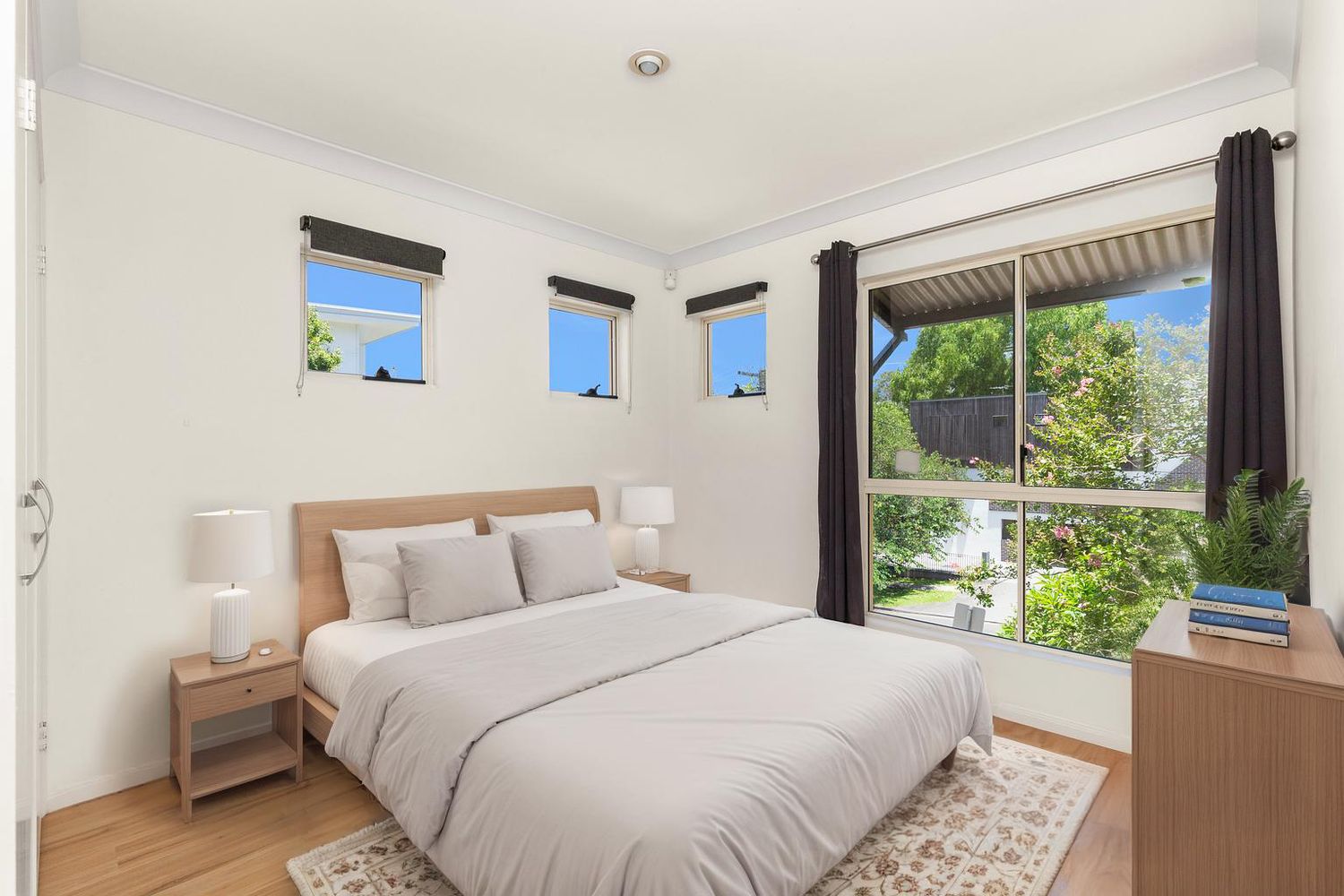 Bright bedroom with queen bed, timber bedside tables, crown molding, and garden views through large window.