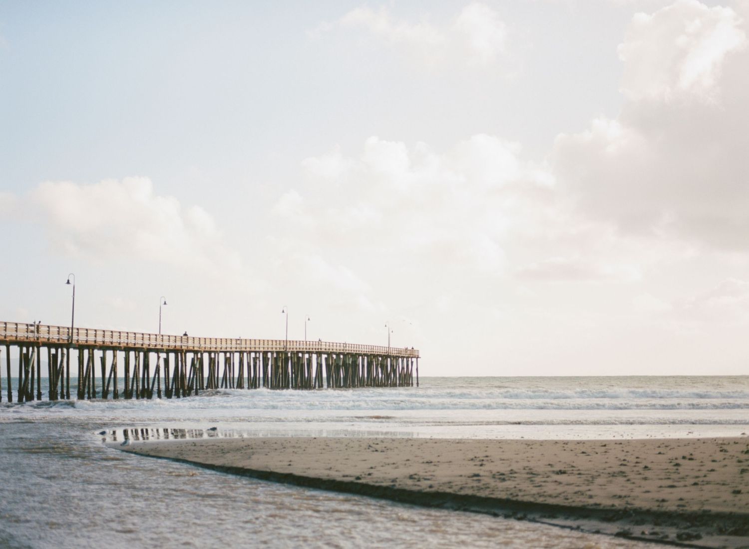 A wooden fishing pier extends into the ocean under a soft cloudy sky at sunset along a sandy beach.