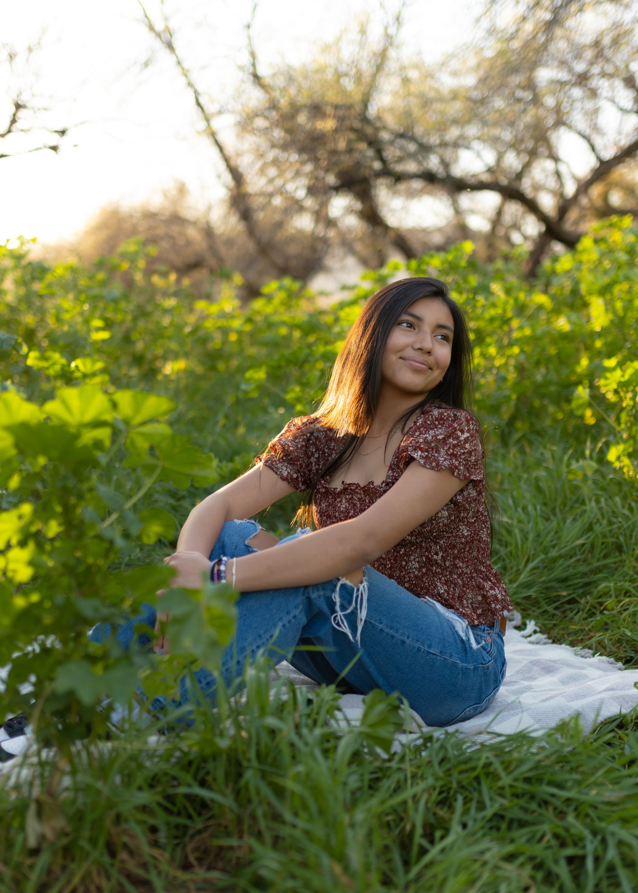 A person in a brown floral top and jeans sits in a field of yellow wildflowers during golden hour.