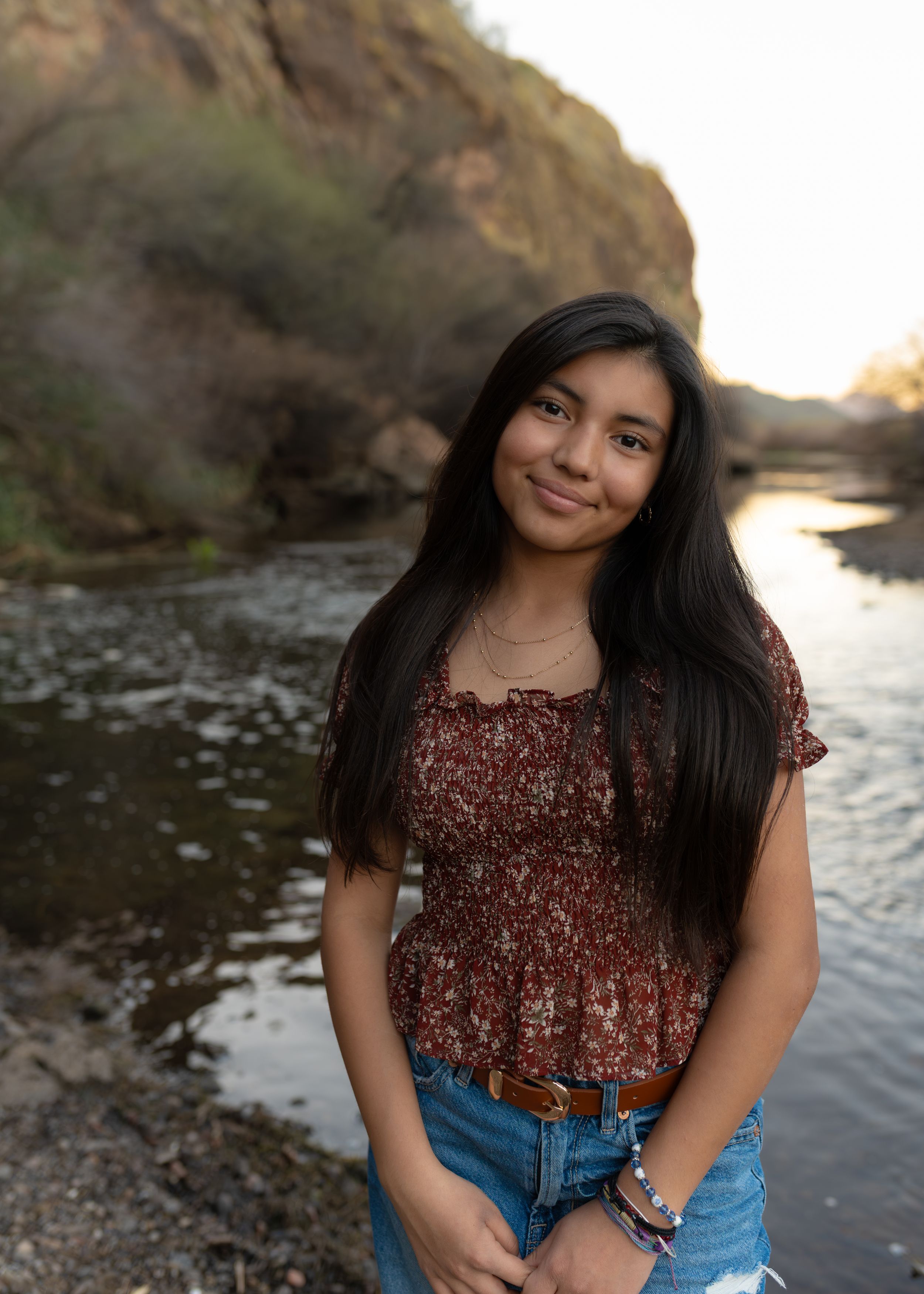 A person in a burgundy sequined top and jeans poses by a riverside at sunset with mountains in the background.