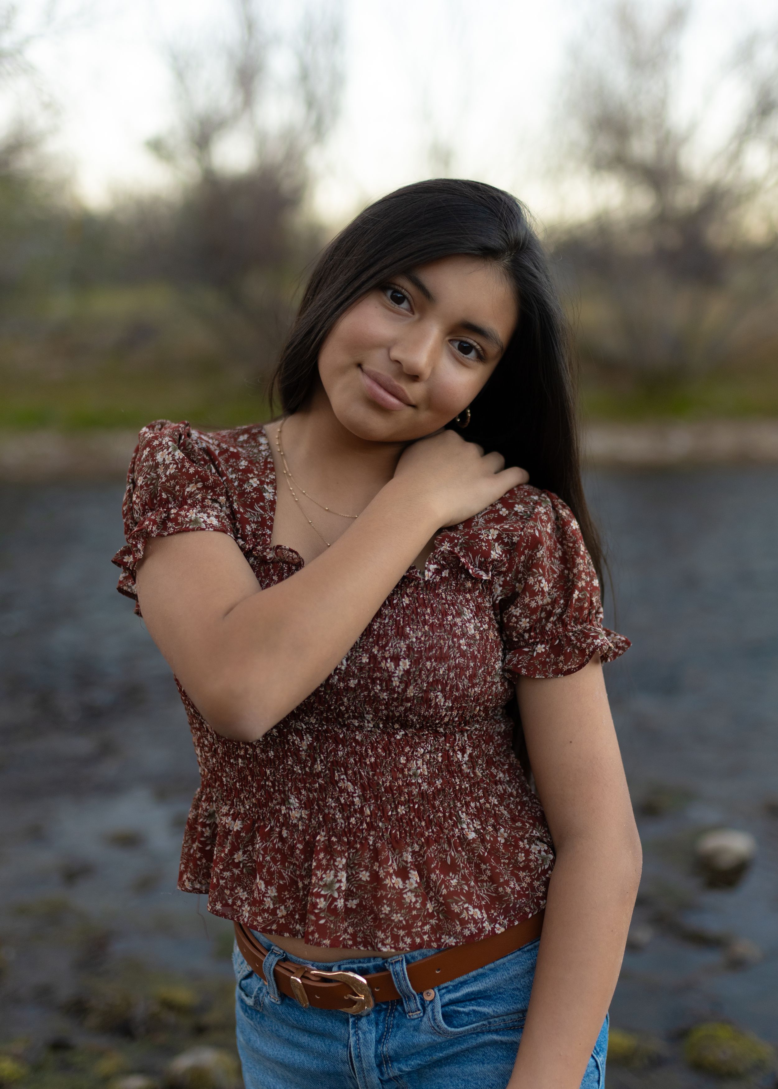 Woman in burgundy floral top and blue jeans poses against blurred outdoor background near water.