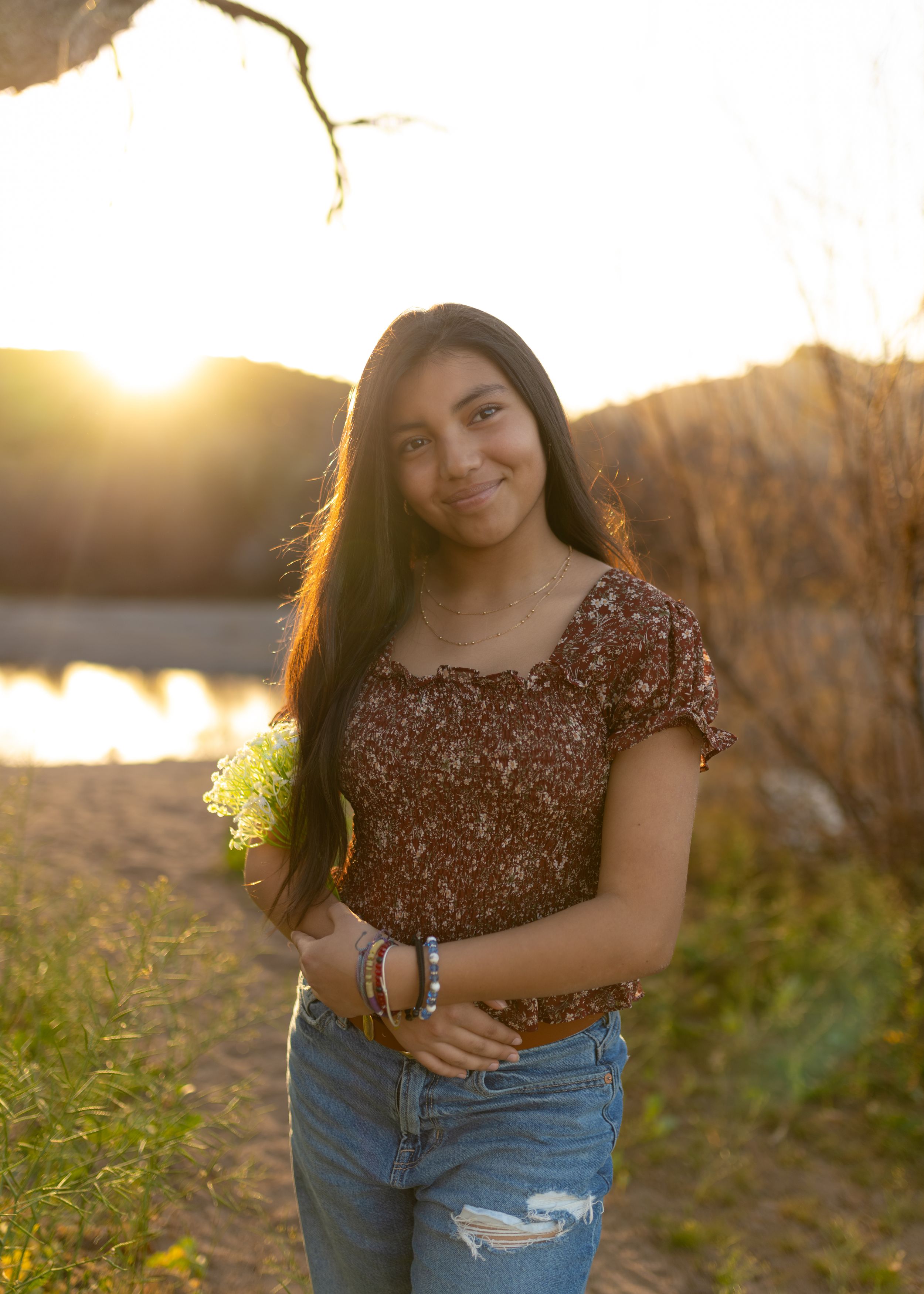 A person in a brown floral top and distressed jeans poses in a golden field at sunset.