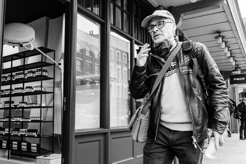 A man in a casual jacket and cap smokes while outside a storefront in black and white.