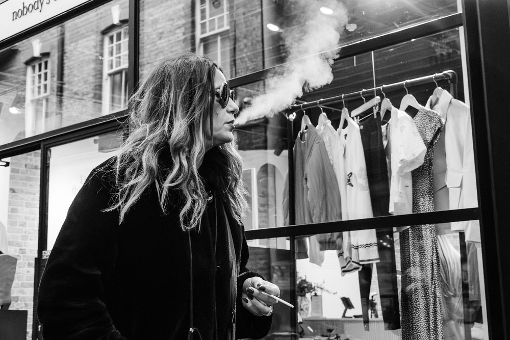 Black and white image of a person smoking while looking at clothing displayed in a storefront window.