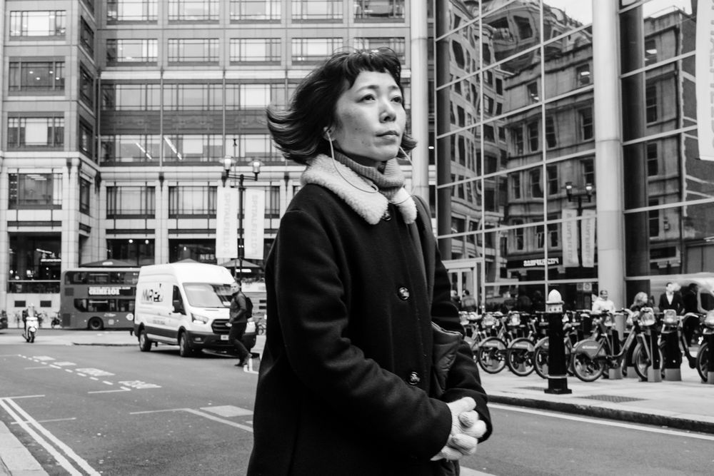 An Asian woman in a black coat walks past modern office buildings on a city street corner. - black & white street photography