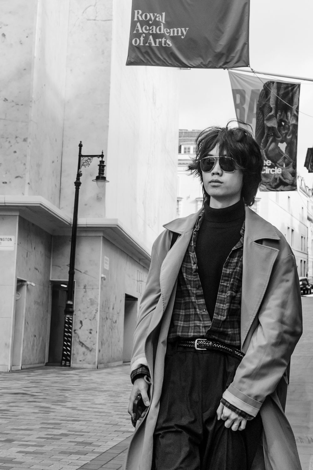 Black and white fashion photo showing person in sunglasses and trench coat outside Royal Academy of Arts building.