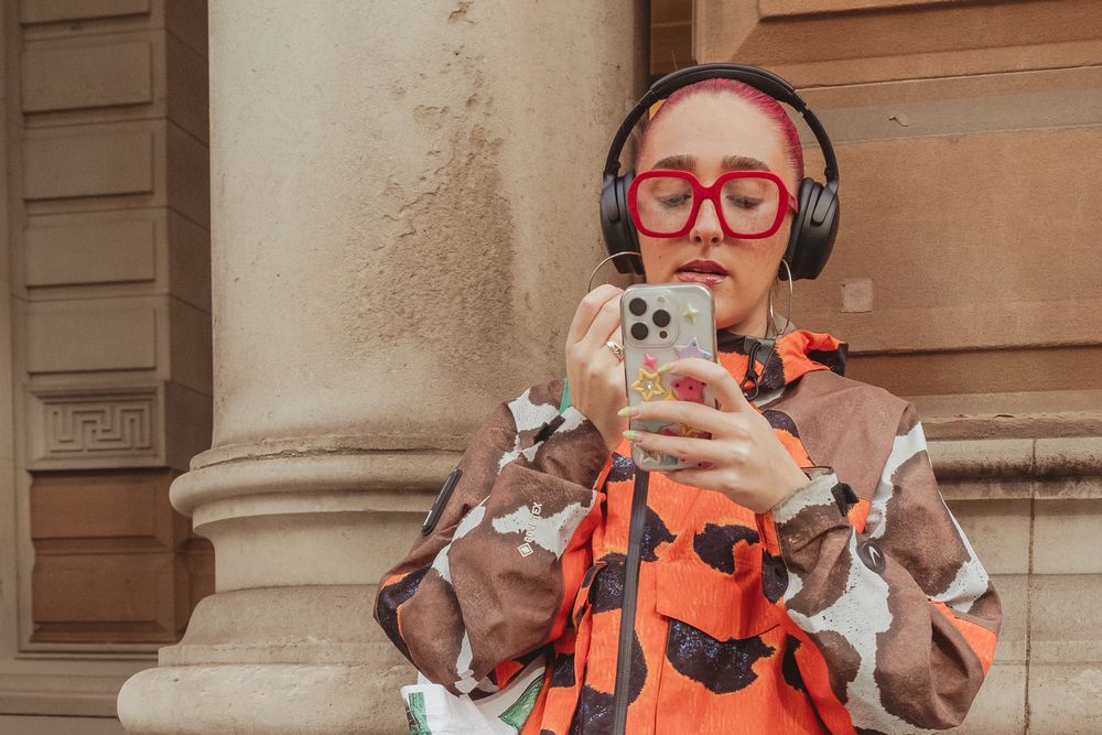 Person in bright orange outfit and red glasses takes selfies while posing against stone building exterior.