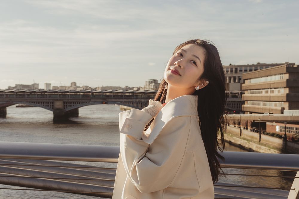 Asian woman in a white sweater poses thoughtfully by a bridge during golden hour with urban architecture in the background.