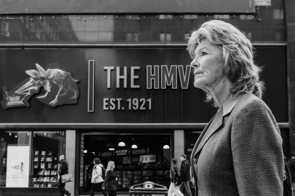 Black and white urban street scene showing people walking past storefronts with a sign reading 'THE HMV' visible.