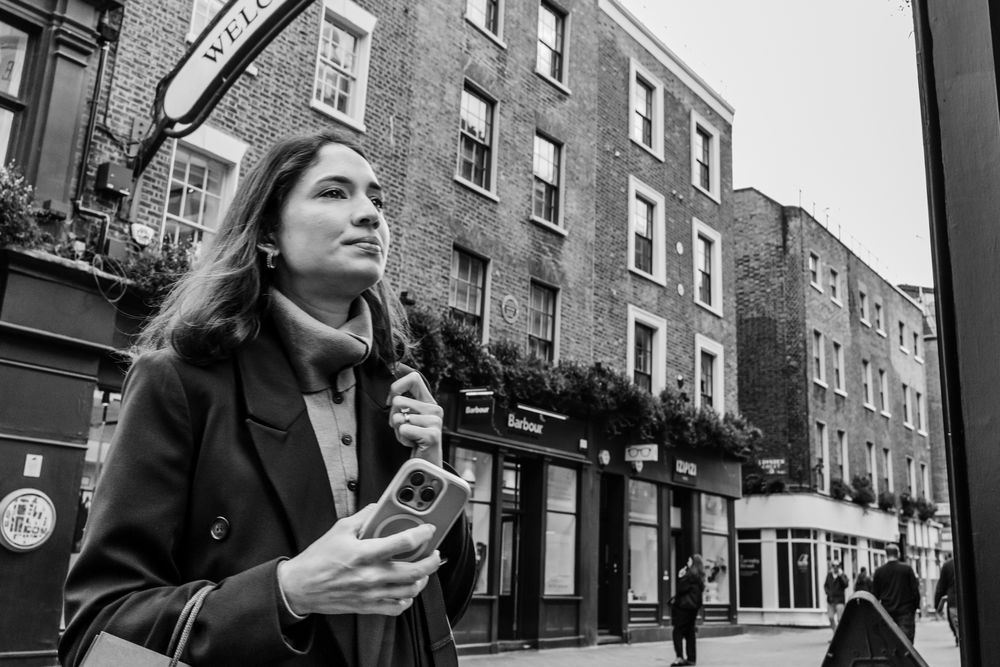 People walking and checking phones on a city street lined with classic brick townhouses.