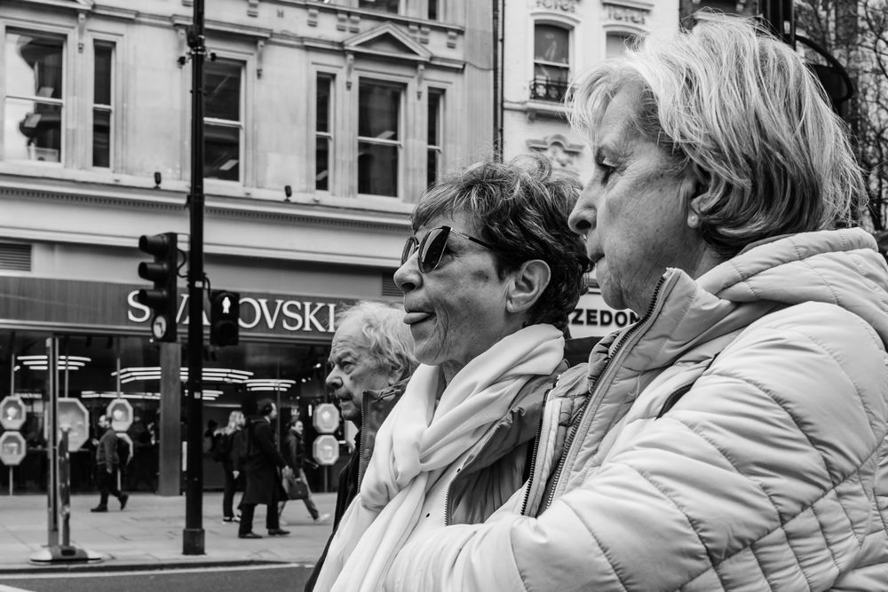 Black and white street photograph showing three people in profile looking to the right with a Swarovski store in the background.