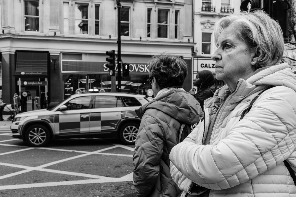A black and white street scene shows pedestrians waiting at a crosswalk while a police car drives past in an urban setting.