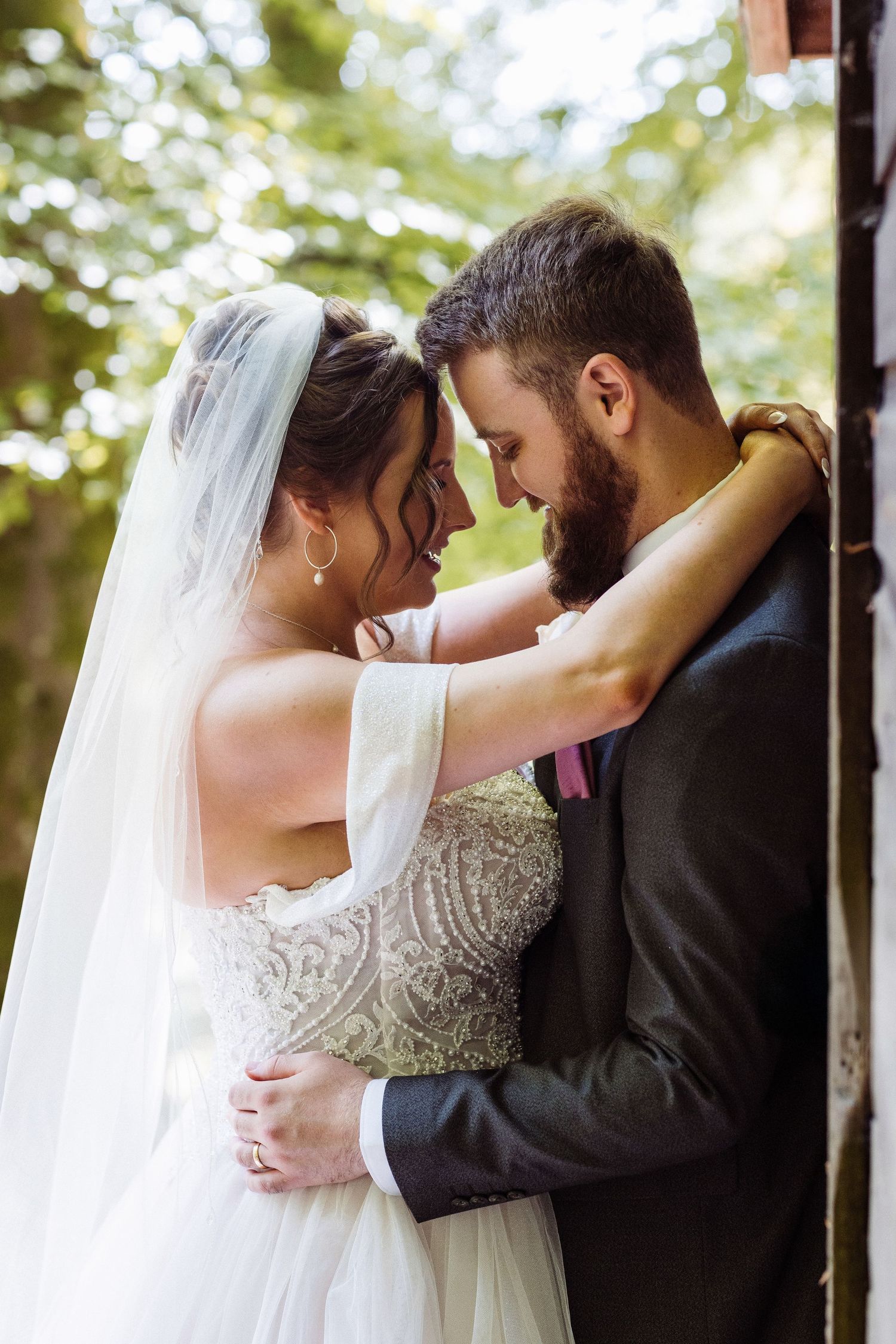 Romantic wedding portrait of newlyweds embracing in soft natural light with flowing white veil and formal attire.