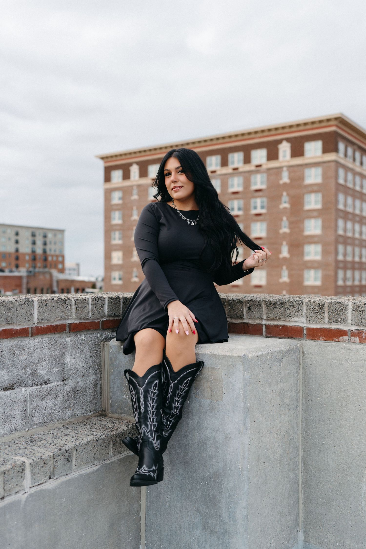 Woman in black attire sits on concrete wall with brick apartment building in background. Portrait Session Wilmington NC