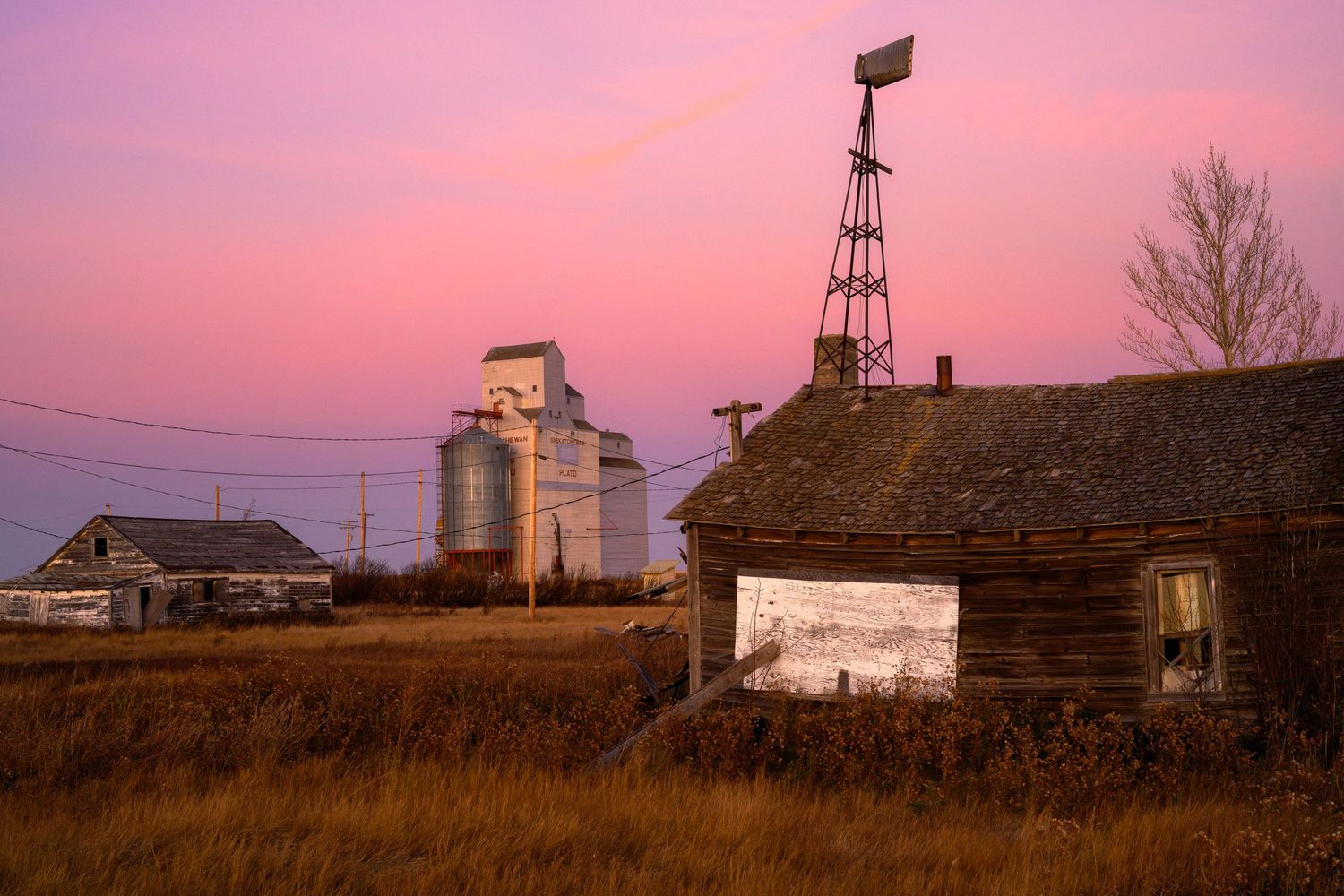 An old wooden farmhouse and grain elevator stand against a pink sunset sky with a windmill silhouetted in rural farmland.