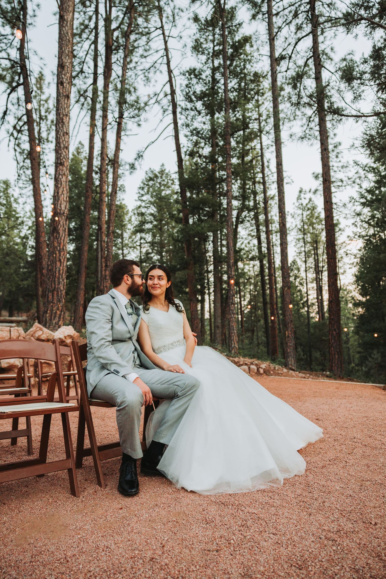 A couple sits together on wooden chairs in a forest setting with tall pine trees and rustic decor at an outdoor wedding.