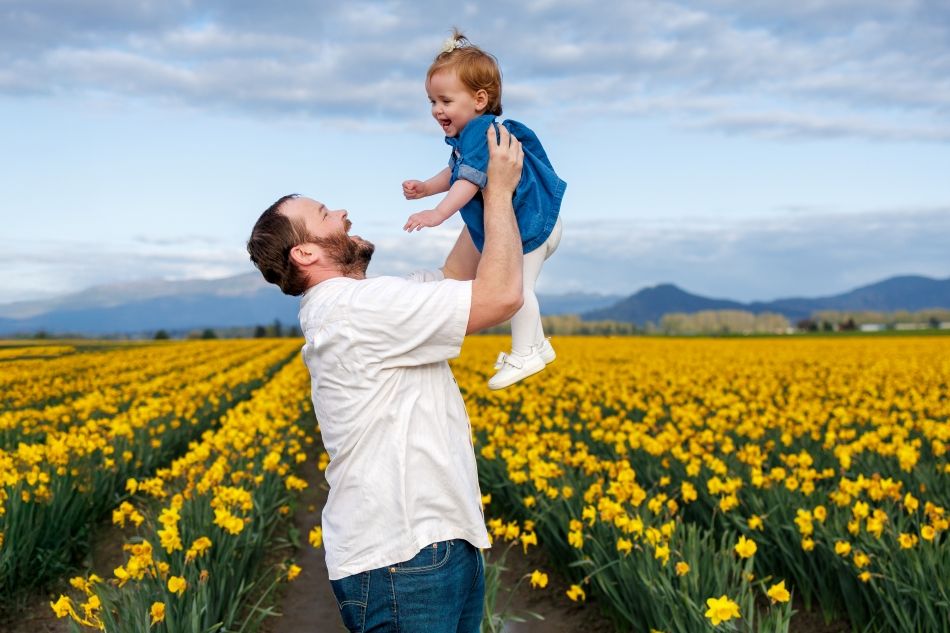 A family plays together in a vibrant yellow daffodil field with mountains in the background on a cloudy spring day.