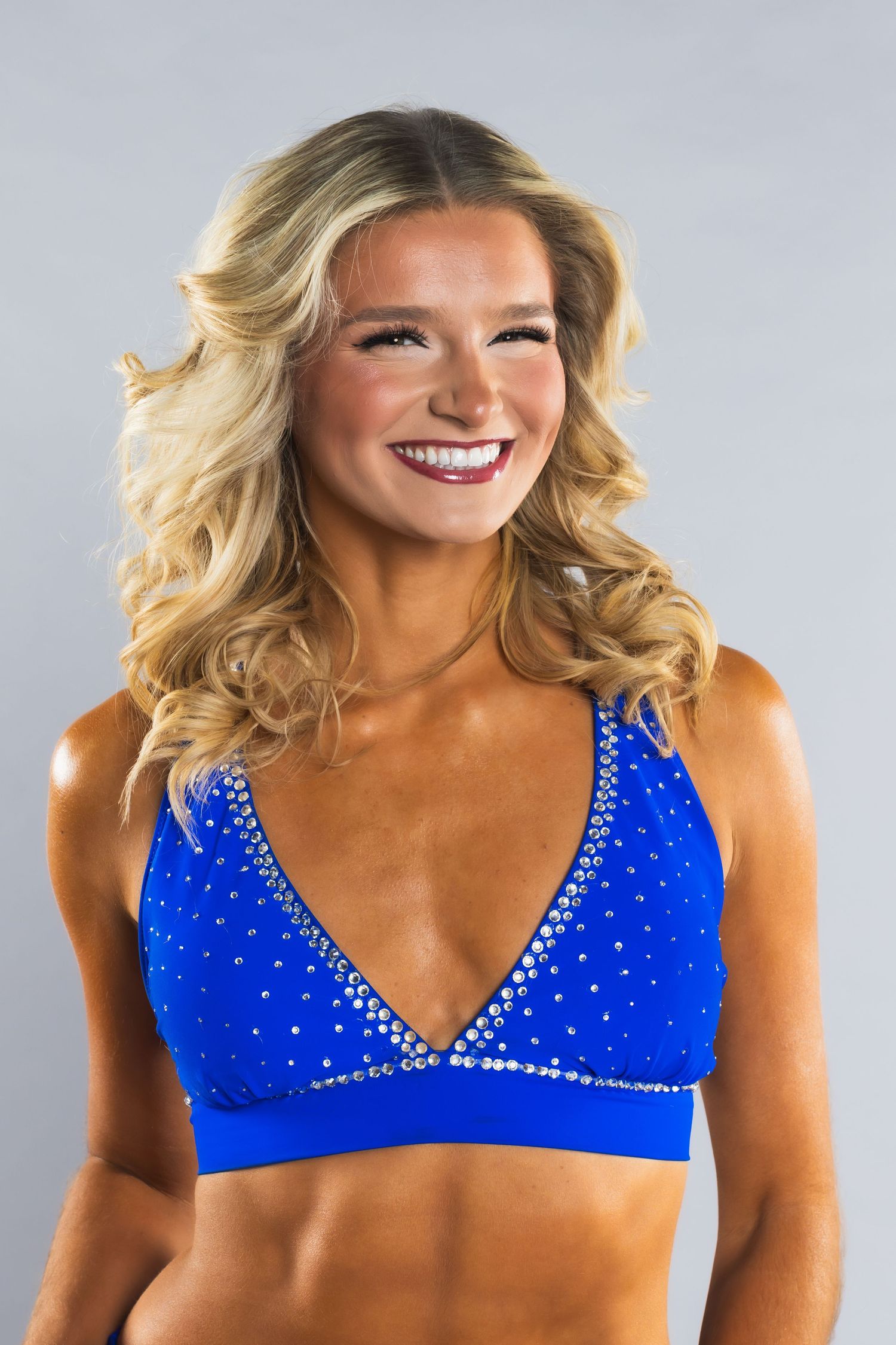 Smiling blonde woman in a blue rhinestone crop top posing confidently against a gray background.