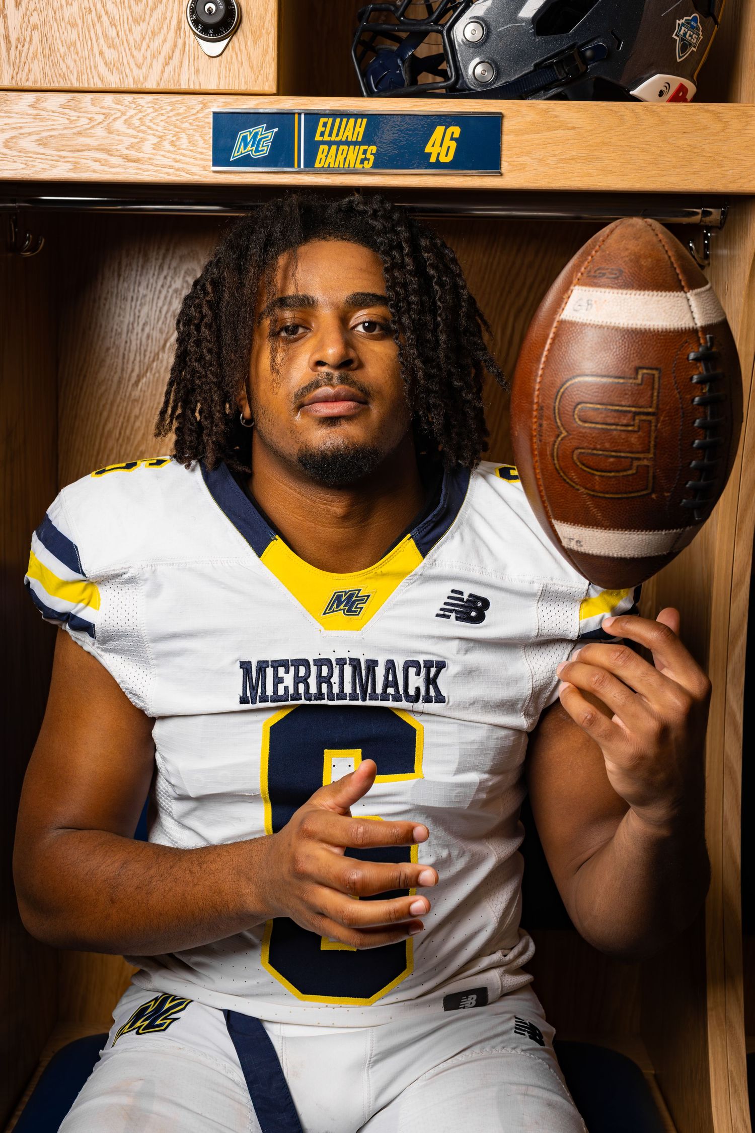 College football player in white Merrimack jersey poses with football in locker room.