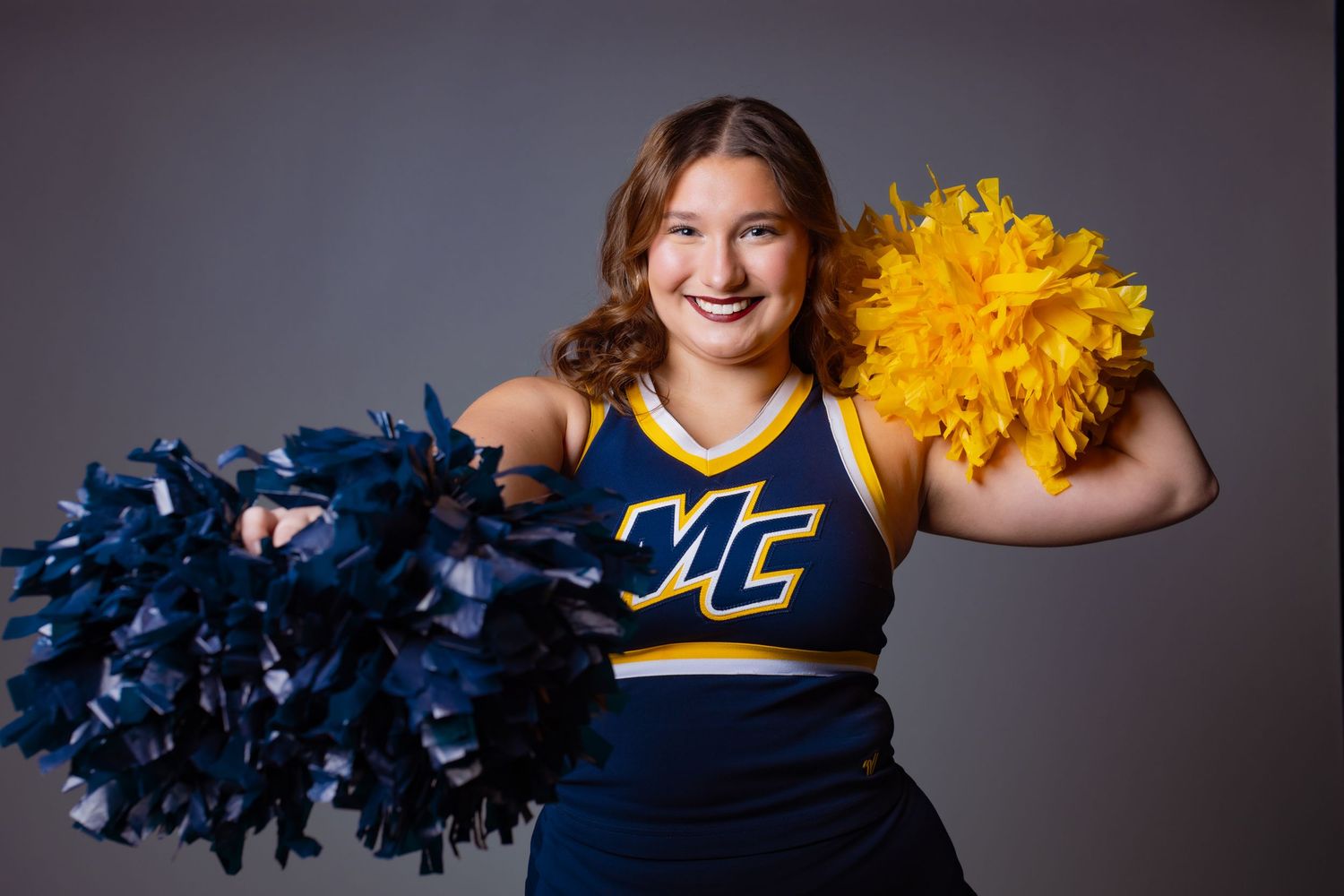 Cheerleader in blue and gold MC uniform smiling with navy and yellow pom-poms in studio portrait.